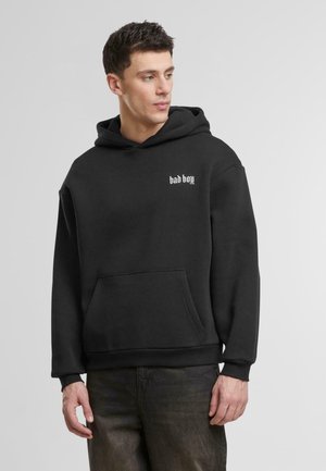 Svart hettegenser med frontlomme, med hvit tekst "bad boy". Stoffet ser mykt og tykt ut, med ribbestrikkede mansjetter og nederkant.