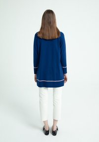 Pullover a maniche lunghe blu navy con bordi in rosa a contrasto, abbinato a pantaloni bianchi a vita alta e calzature nere, mostrato da dietro.