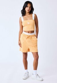 Oranje crop top met vierkante halslijn en witte geborduurde merklogo, gecombineerd met elastische shorts met zijzakken in een vergelijkbare kleur.