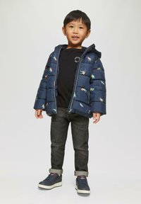Jonge jongen in een marineblauwe pufferjack met dinosauruspdruk, zwart T-shirt, opgerolde donkere spijkerbroek en marineblauwe sneakers, staand tegen een effen achtergrond.