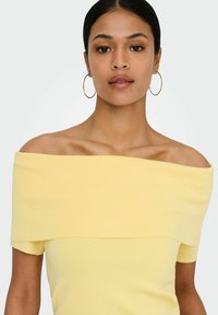 Jeune femme avec un chignon lisse portant des boucles d'oreilles créoles dorées et un haut jaune pâle à épaules dénudées et côtelé, regardant vers l'avant sur un fond blanc.