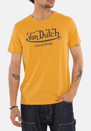 Homme portant un t-shirt jaune moutarde Von Dutch California avec des tatouages sur les deux avant-bras et un jean bleu foncé, main dans la poche.
