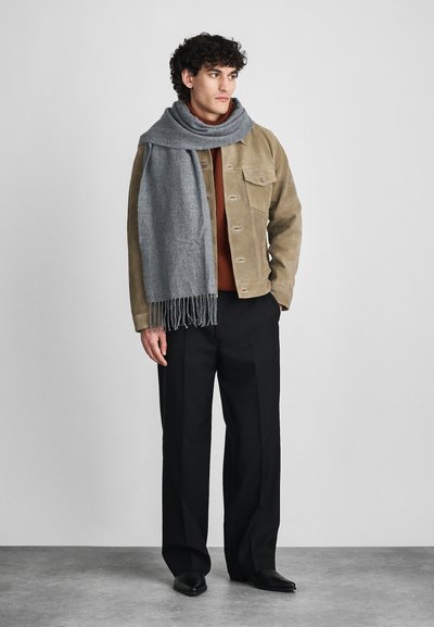 Μπεζ βελούδινο σακάκι, καφέ πουλόβερ, γκρι κασκόλ με φούντες, μαύρο παντελόνι και παπούτσια. Το μοντέλο στέκεται μπροστά από έναν απλό γκρι φόντο.