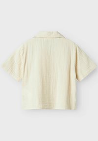 Camisa de manga corta de color beige claro con un acabado texturizado, que presenta un collar sencillo y un corte rectangular. Sin patrones ni herrajes visibles.