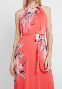 Robe halter en corail avec imprimé floral rose et vert. Dotée d'une ceinture à la taille et d'une texture de tissu fluide. Design sans manches.