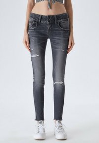 Jeans ajustados en denim negro desgastado, con roturas y detalles desgastados, con un dobladillo sin terminar. Combinados con zapatillas blancas.
