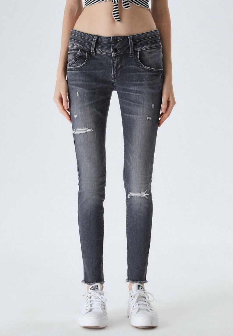 Jeans ajustados en denim negro desgastado, con roturas y detalles desgastados, con un dobladillo sin terminar. Combinados con zapatillas blancas.