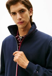 Giacca zip-up blu marina con colletto alto, caratterizzata da una cerniera rossa e bianca. Sotto, è visibile una maglietta a righe bordeaux e bianche.