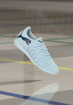 Lichtblauwe Puma zaalvoetbalschoen met zwart logo, veters gestrikt, zwevend boven een sportvloergedeelte met lijnen van het speelveld.