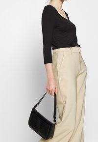 Nainen, jolla on päällään musta pitkähihainen paita ja vaaleanbeiget korkeavyötäröiset housut, pitää kädessään pientä mustaa käsilaukkua, jossa on lyhyt olkahihna.