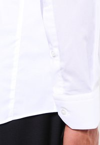 Witte overhemd mouw met dubbele knoopmanchet, gedragen met donkere gestructureerde broek.