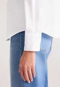 Camicia bianca con bottoni e dettagli ai polsini, abbinata a jeans in denim blu. La camicia presenta una texture liscia e una lucentezza sottile.