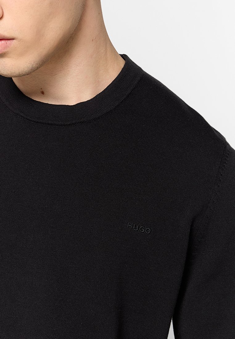 Schwarzer Strickpullover mit rundem Ausschnitt, der ein dezentes eingeprägtes Logo auf der Brust hat. Glatte Textur, figurbetontes Design und lange Ärmel.