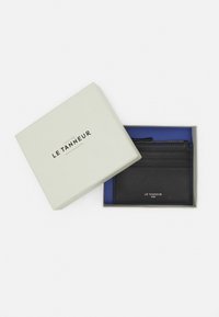 Porte-cartes en cuir noir avec finition texturée, coutures bleues et plusieurs emplacements, présenté dans une boîte vert clair avec un intérieur bleu.