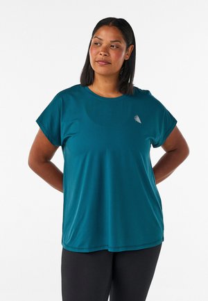 Frau mit dunklen Haaren, die ein türkisfarbenes Kurzarmshirt mit kleinem weißen Logo trägt, steht mit den Händen hinter dem Rücken und schaut leicht nach rechts.