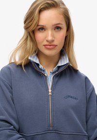 Sweatshirt zippé bleu marine avec une fermeture éclair dorée, présentant un logo brodé discret et porté par-dessus un col de chemise rayé bleu clair.