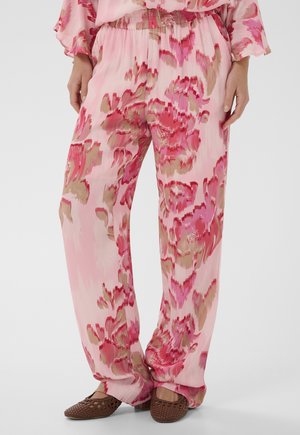 Kobieta ubrana w luźne, różowe spodnie w kwiatowy wzór oraz brązowe, plecione buty zakryte na palce, stojąca z rękami swobodnie ułożonymi wzdłuż ciała.