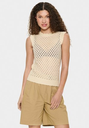 Pulover tricotat fără mâneci, de culoare bej deschis, cu un model texturat și brodat. Decupaj cu margine ondulată, asortat cu pantaloni scurți lejeri de culoare khaki.