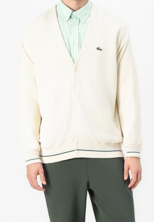 Mann trägt cremefarbenen, geknöpften Cardigan mit kleinem grünem Krokodil-Logo über grün gestreiftem Hemd und dunkelgrüner Hose.