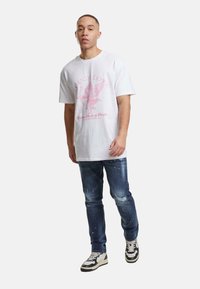 Joven con camiseta gráfica blanca, jeans desgastados azules y zapatillas en blanco y negro caminando hacia adelante contra un fondo liso.