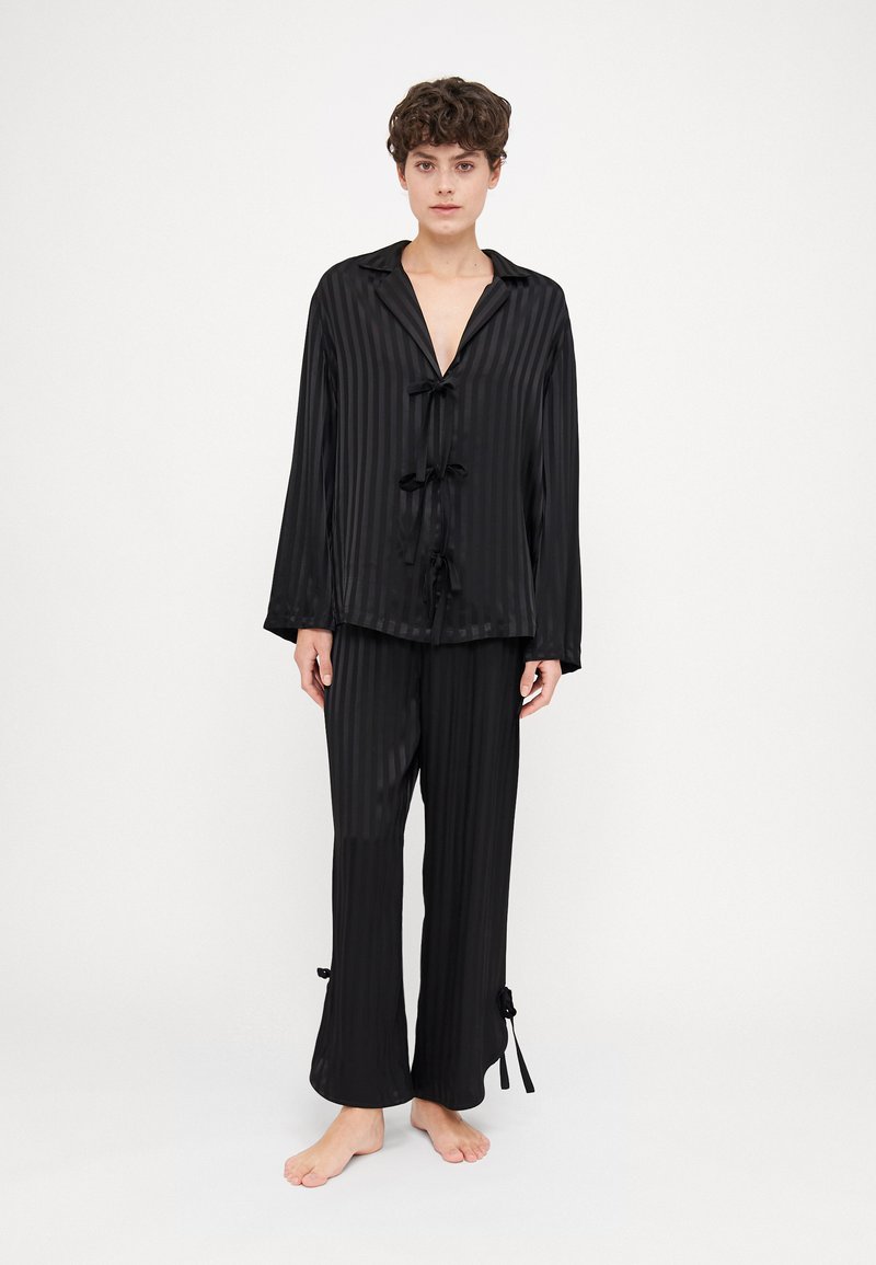Conjunto de pijama de satén negro con rayas verticales; la parte superior cuenta con un collar y lazos en el frente, mientras que los pantalones tienen puños atados. Textura suave y ajuste relajado.