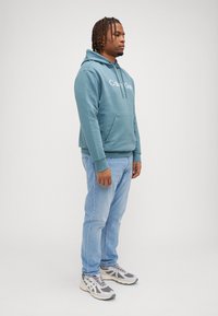 Sweatshirt capuz verde-água com bolso frontal e logótipo branco, combinado com jeans azul-claro e sapatilhas cinzas. Fundo neutro.