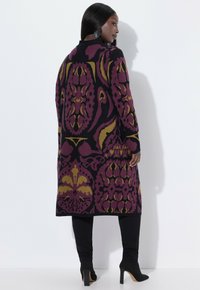 Knielanger, gemusterter Cardigan in tiefem Lila und Gold, mit kunstvollen Designs, langen Ärmeln und einer taillierten Silhouette, kombiniert mit schwarzen Hosen.