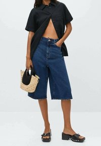 Camicia nera a maniche corte, culotte in denim blu a vita alta, borsa di paglia intrecciata con manici neri e sandali neri con plateau e borchie.