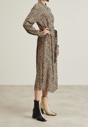 Vrouw die een lange, ceintuurbare jurk met luipaardprint draagt en ongelijkwaardige zwarte en beige hoge-gehakte enkellaarsjes, staand op een neutrale vloer.