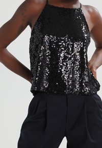 Haut à sequins noirs avec un dos nu, coupe ample et surface texturée, avec un col montant et de fines bretelles. Porté avec un pantalon sombre.