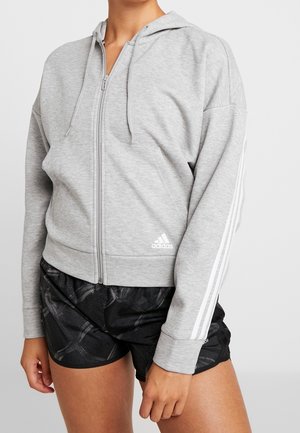 Personne portant un sweat à capuche zippé gris clair avec des rayures blanches sur les manches et un short de sport noir avec un motif subtil.