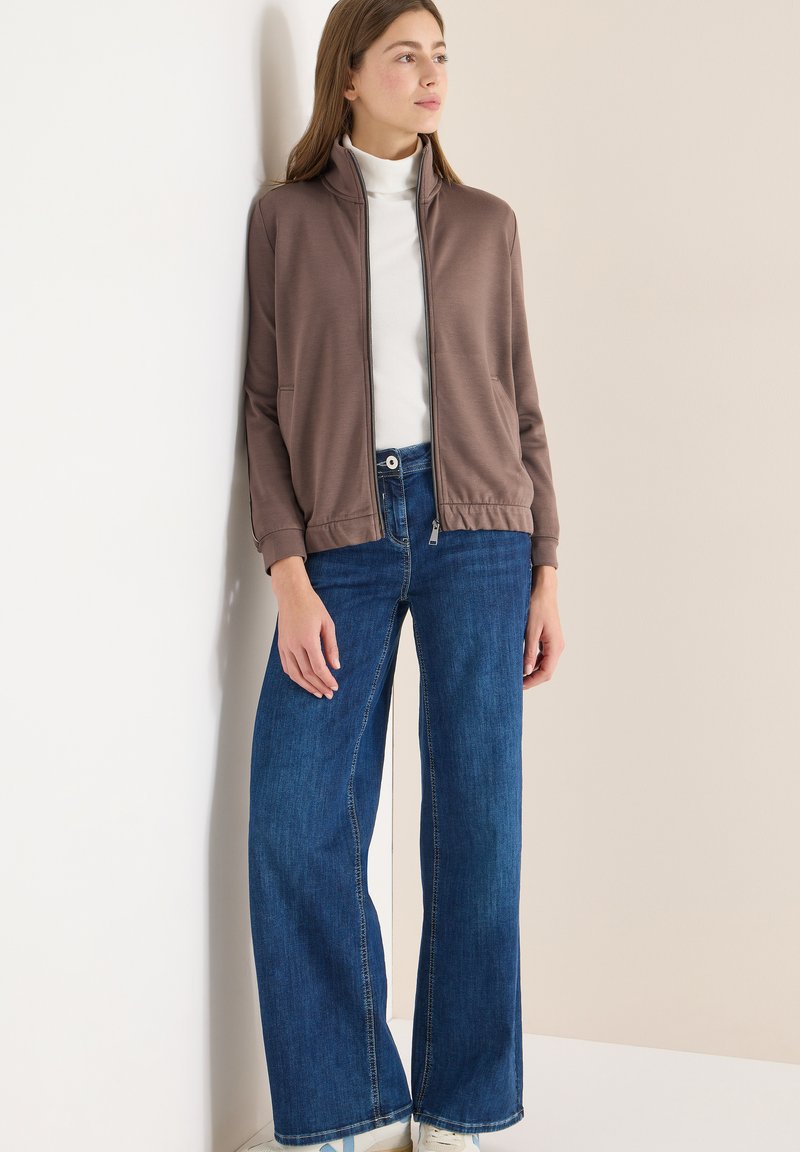 Giacca marrone con zip e collo alto, abbinata a jeans blu a gamba larga. Tessuto dalla texture liscia e vestibilità casual e rilassata.