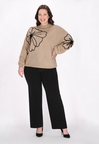 Pull en tricot beige avec broderie florale noire sur les manches, associé à un pantalon noir et des escarpins noirs. La texture est douce et ample.