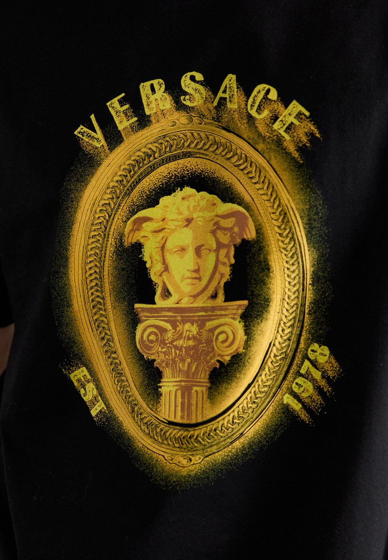 Geel Medusa-hoofd en sierlijke kolomontwerp met "VERSACE" erboven en "EST 1978" eronder op een zwarte stoffen achtergrond.