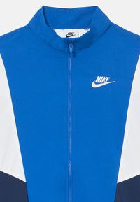Nike modrá bežecká bunda s vysokým golierom, zipsom vpredu a kontrastnými bielymi a námorníckymi panelmi. Hladká, ľahká tkanina.