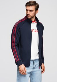 Marineblaues Zip-Up-Jacket mit roten Seitenstreifen und dem Text "Performance"; getragen über einem weißen T-Shirt und kombiniert mit hellblauen Jeans.