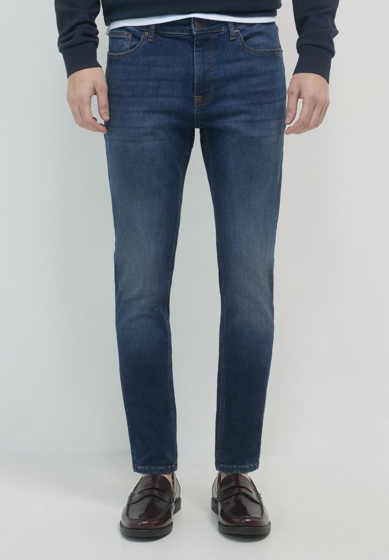 Uomo che indossa jeans slim-fit blu scuro e scarpe slip-on in pelle marrone scuro, in piedi contro uno sfondo chiaro e semplice.