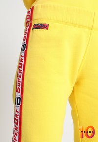 Geel joggingbroek gemaakt van zachte stof, met een grote rood-witte logo-streep aan de zijkant en een klein merklabel op de heup.