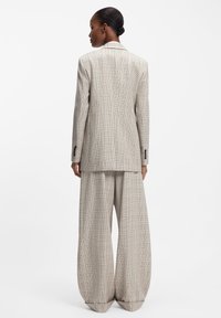 Rudebenet skræddersyet jakkesæt i beige med lysebrune og blå accenter. Løse bukser supplerer en struktureret blazer med hakket krave.