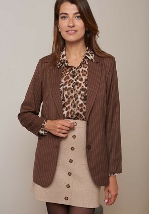 Blazer marron à rayures, porté sur un blouse à imprimé léopard, associé à une jupe beige à boutons. Textures lisses et coupe ajustée.