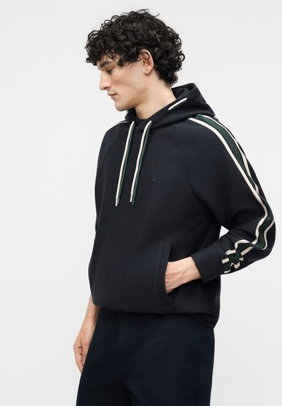 Νέος άνδρας με σγουρά μαλλιά που φοράει μαύρο φούτερ με κουκούλα με πράσινες και λευκές ρίγες στα μανίκια, με ένα χέρι στην τσέπη, κοιτάζοντας προς τα αριστερά μπροστά σε λευκό φόντο.