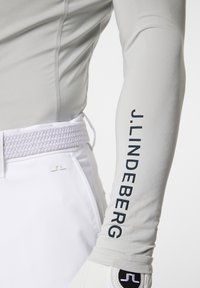 Maglietta a maniche lunghe grigio chiaro con dettagli sulle cuciture laterali e logo "J.LINDEBERG" stampato in blu scuro sul braccio. Abbinate a una cintura bianca e guanti.