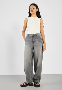 Weißes, tailliertes, ärmelloses Top, kombiniert mit hellgrauen, weiten Jeans. Die Jeans haben eine hohe Taille und eine dezente Streifenstruktur. Schwarze Plateausandalen.
