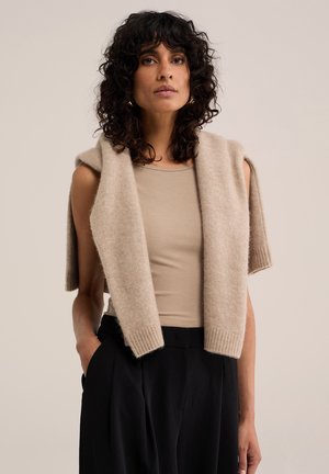 Frau mit lockigem dunklem Haar trägt ein beiges ärmelloses Top und einen beigen Pullover über den Schultern sowie schwarze Hose, steht vor neutralem Hintergrund.