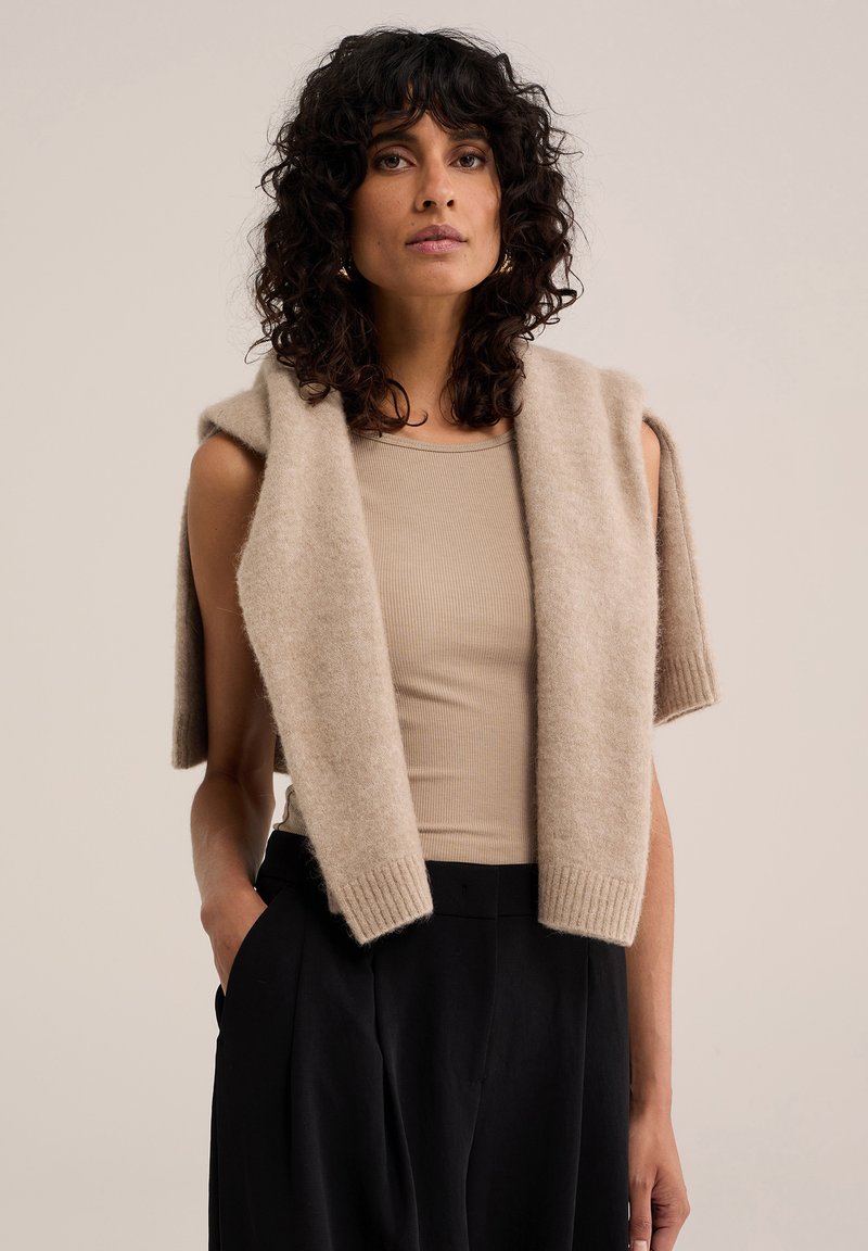 Femme aux cheveux bouclés et foncés porte un top beige sans manches et un pull beige posé sur ses épaules avec un pantalon noir, debout devant un fond uni.
