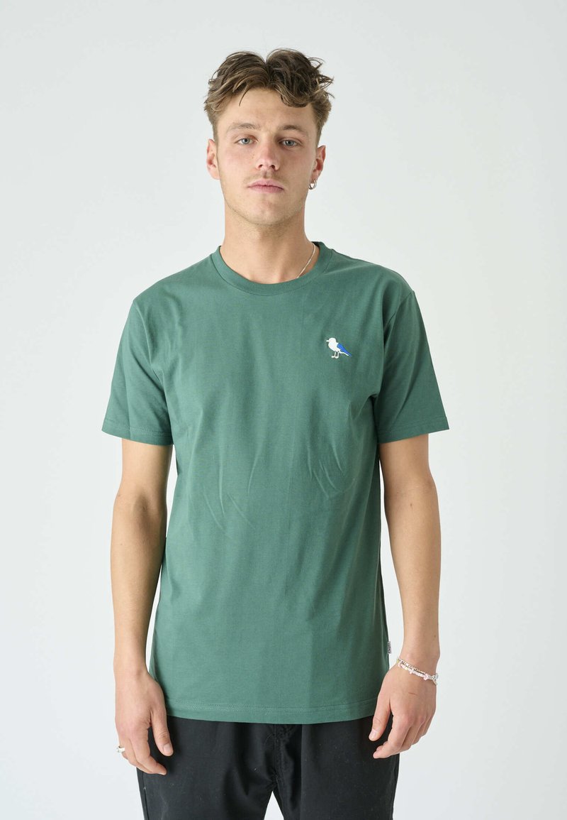 Grünes Baumwoll-T-Shirt mit Rundhalsausschnitt, kurzen Ärmeln und einer kleinen blau-weißen Vogelstickerei auf der linken Brust.