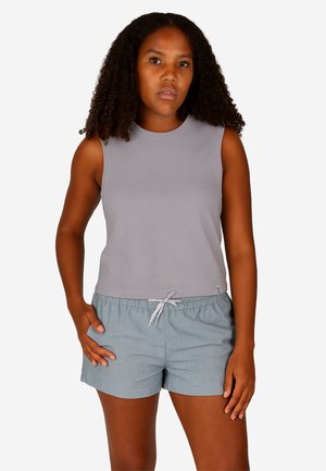Woman with curly hair wearing a sleeveless gray top and light blue shorts with a white drawstring, standing with one hand in pocket.