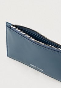 Μπλε δερμάτινο πορτοφόλι με φερμουάρ, με λείο υφή και λευκό λογότυπο. Το εσωτερικό είναι ορατό, αποκαλύπτοντας μια αντίθετη επένδυση.