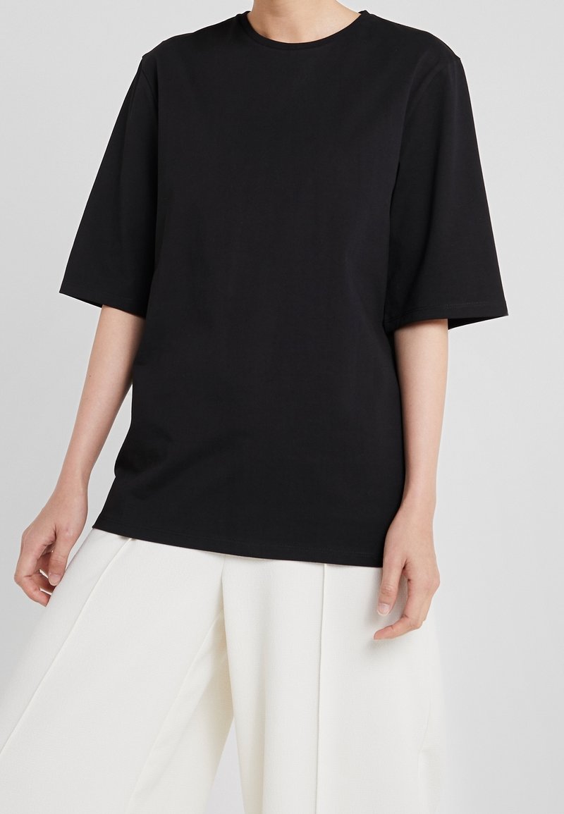 Černé bavlněné tričko s kulatým výstřihem a krátkými rukávy, s volnějším střihem a hladkou texturou. V kombinaci s bílými kalhotami s širokými nohavicemi.