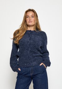 Tumši zila adīta kardigāna ar strukturētu apdari, ar apaļu kakla izgriezumu, pogu aizdari un garām piedurknēm. Valkāts ar tumšiem džinsiem.